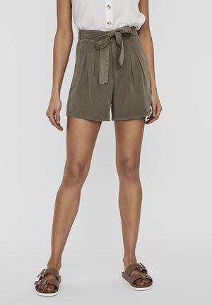 Shorts kaki avec des plis, taille haute, et une ceinture assortie. Associés à des sandales à glisser marron, avec des accents métalliques.