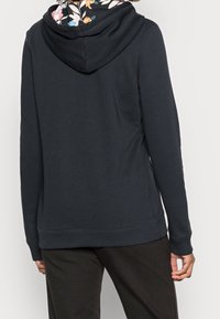 Persona vestida con una sudadera negra con un interior de capucha con estampado floral, de espaldas, combinada con pantalones negros, contra un fondo liso.