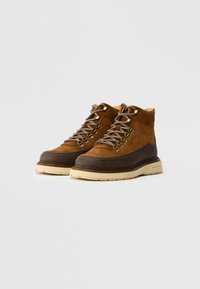 Braune Wildleder-Stiefeletten mit einer dunkelbraunen Gummisohle, ausgestattet mit metallischen Ösen, grauen Schnürsenkeln und einer cremefarbenen Sohle.