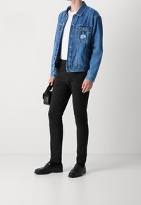 Džínová bunda středně modré barvy, bílé tričko, černé slim-fit kalhoty a černé mokasíny, držící malou černou tašku. Jednoduchý, moderní design.