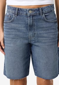 Gaiši zilas džinsu šorti ar augstu jostasvietu, piecu kabatu dizainu un saplēstām malām. Materiāls šķiet teksturēts ar klasisku džinsas apdari.