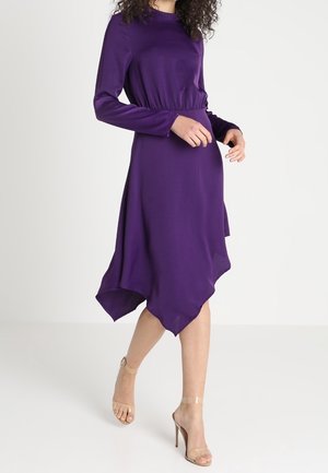 Femme marchant dans une robe violette à manches longues avec un ourlet asymétrique, portant des sandales à talons hauts avec des brides transparentes.