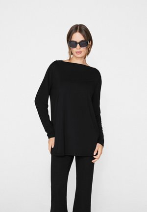 Vrouw die een oversized zwarte top met lange mouwen en broek draagt, met grote zwarte zonnebril, staand tegen een effen witte achtergrond.