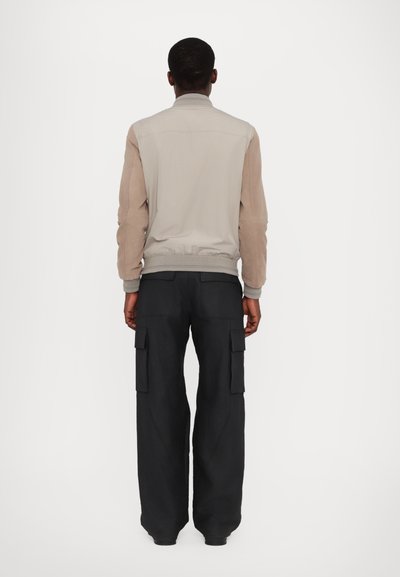 Helle beige Bomberjacke mit einer strukturierten braunen Oberseite, geripptem Kragen und dunklen Cargo-Hosen mit mehreren Taschen.