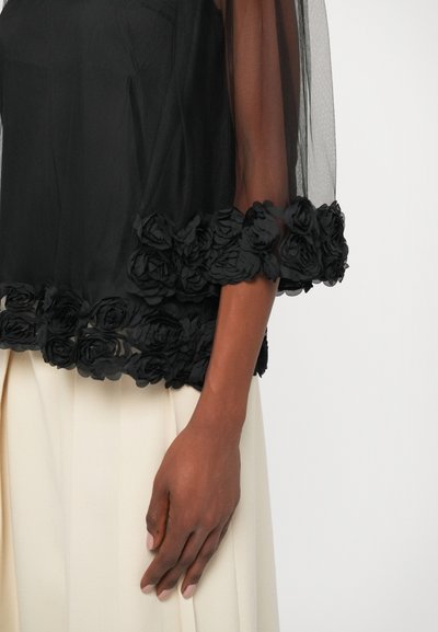Black sheer top with floral appliqué at the hem, featuring a loose fit and semi-transparent sleeves, paired with a pleated cream skirt.