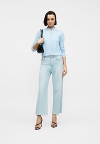 Femme portant un cardigan boutonné bleu clair et un jean large délavé, tenant un sac à main noir, debout sur un fond blanc.