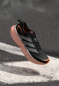 Scarpa sportiva nera con tre strisce argento, accenti arancioni, tomaia in mesh strutturato e suola leggera rosa, mostrata a mezz'aria.