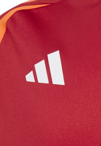 Sfondo tessile rosso con un accento arancione. Presenta un logo a tre strisce bianche prominente, tessuto testurizzato e superficie liscia.