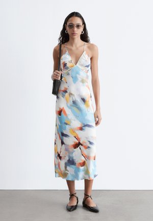 Femme portant des lunettes de soleil, des boucles d'oreilles créoles, une robe nuisette fleurie colorée, des ballerines noires à lacets à la cheville, et portant un petit sac à épaule noir, debout.