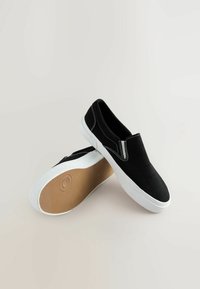 Schwarze Slipper mit weißer Sohle, ausgestattet mit kontrastierenden weißen Nähten und elastischen Seiten. Glattes Material mit einer strukturierten Gummisohle.