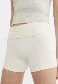 Kobieta w obcisłych, beżowych krótkich spodenkach sportowych z wysokim stanem i jasnoszarej, skróconej koszulce na tle białego tła.
