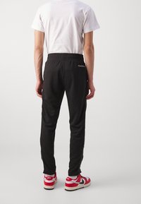 Zwarte sportbroek met taps toelopend ontwerp, elastische taille, zijzakken en contrasterende ritsen. Draagt rode en witte sneakers.