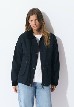 Veste bleu marine avec fermeture éclair, col, et deux poches avant. Le tissu semble lisse avec un léger éclat. Assortie à un pull blanc.