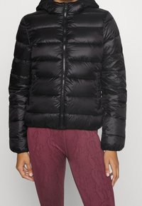 Svart puffjacka med huva, med kviltad design, slätt nylonmaterial och en dragkedja i fronten. Bärs med texturerade bordeaux leggings.