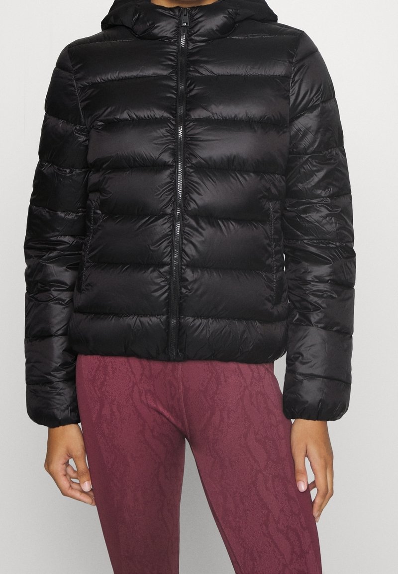Svart puffjacka med huva, med kviltad design, slätt nylonmaterial och en dragkedja i fronten. Bärs med texturerade bordeaux leggings.