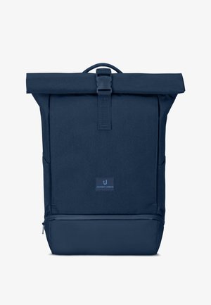 Junge Frau trägt einen dunkelblauen Roll-Top-Rucksack mit Reißverschlusstasche, trägt ein schwarzes T-Shirt und blaue Jeans, steht vor einem weißen Hintergrund.