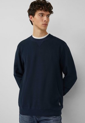 Mann mit lockigem Haar, der einen marineblauen Rundhalsausschnitt-Pullover über einem weißen Hemd trägt und mit den Händen hinter dem Rücken vor einem schlichten grauen Hintergrund steht.