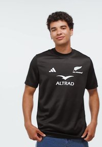 Nuori mies, joka pukeutunut mustaan All Blacks -rugby-paitaan, jossa on Adidas- ja Altrad-logot. Hän pitää paidan helmaa kiinni ja seisoo yksinkertaista valkoista taustaa vasten.