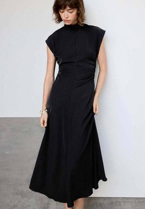 Mujer con cabello corto y castaño que lleva un vestido negro midi sin mangas, de pie frente a un fondo liso blanco y gris.