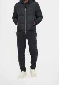 Veste matelassée noire avec capuche, dotée d'une fermeture éclair frontale et de poignets élastiques ; associée à un pantalon de survêtement noir et des baskets de couleur claire.