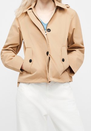 Kvinde iført en beige jakke med sorte knapper og lynlås over en blå top, kombineret med hvide bukser, hænderne i jakkelommerne.