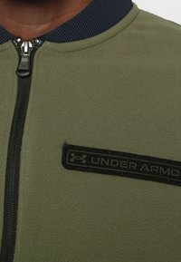 Olivově zelená fleecová bunda s tmavě modrým žebrovaným límcem, černým zipem a nášivkou s logem Under Armour na levé straně hrudi. Měkká textura.