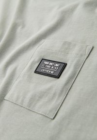 Camiseta de algodón verde claro con un bolsillo de parche que presenta una etiqueta rectangular negra. Texto en la etiqueta: "SU ÚNICO LÍMITE ES SU MENTE" y "Ombre."