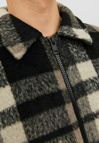 Giacca in pile a quadri con motivo check in nero, grigio e crema. Presenta una zip completa e un colletto, mettendo in mostra un materiale morbido e testurizzato.