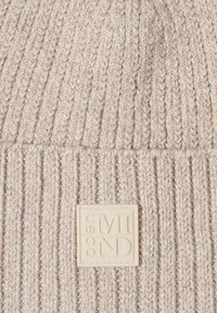 Gorro de lana en beige claro, con textura acanalada, que presenta un parche cuadrado prominente con "open MIND" en relieve en crema. Material suave.