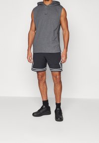 Canottiera grigia senza maniche con tasca frontale, abbinata a pantaloni corti neri con strisce bianche laterali. Sneakers nere completano il look sportivo.