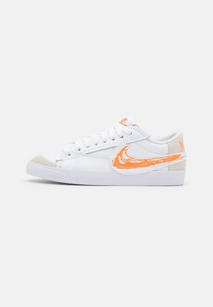 Valkoiset matalavartiset tennarit, joissa on oranssi siveltimellä tehty Nike-logon kuvio, valkoiset nauhat, beige mokkanahkainen kärki ja tasainen valkoinen kumipohja.