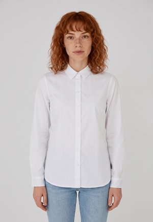 Vrouw met krullend rood haar die een witte blouse met knopen en lichtblauwe spijkerbroek draagt, staand tegen een effen lichtgrijze achtergrond.
