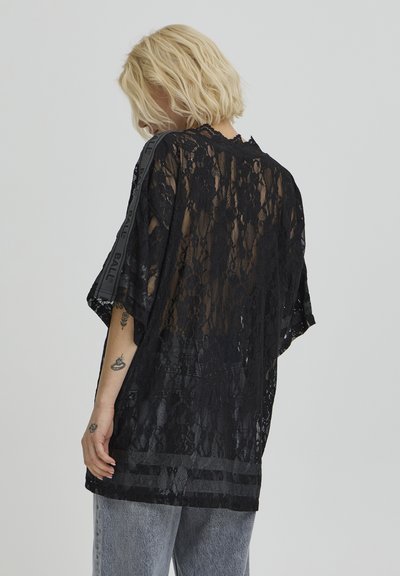 Haut en dentelle noire à manches courtes, présentant une texture transparente, un ourlet incurvé et des accents à rayures verticales le long des épaules.