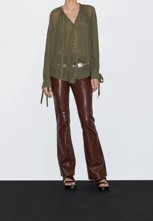 Femme portant un chemisier vert olive transparent avec des détails de boutons et un pantalon évasé en cuir marron, associé à des sandales à plateforme noires.
