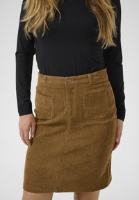 Brun corduroybukserkirt med to frontlommer, en knaplukning i denim-stil og et lige design. Parret med en langærmet sort top.