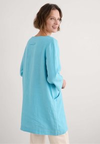 Lichtblauwe tuniek van linnen, met lange mouwen, een ronde halslijn en zijzakken. De achterkant heeft een klein, rechthoekig detail.