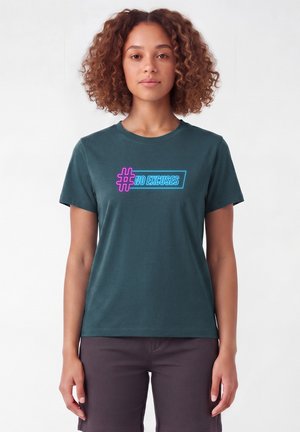 Vrouw met krullend haar die een donker turquoise shirt draagt met neonroze en blauwe tekst "# NO EXCUSES", staand tegen een effen witte achtergrond.