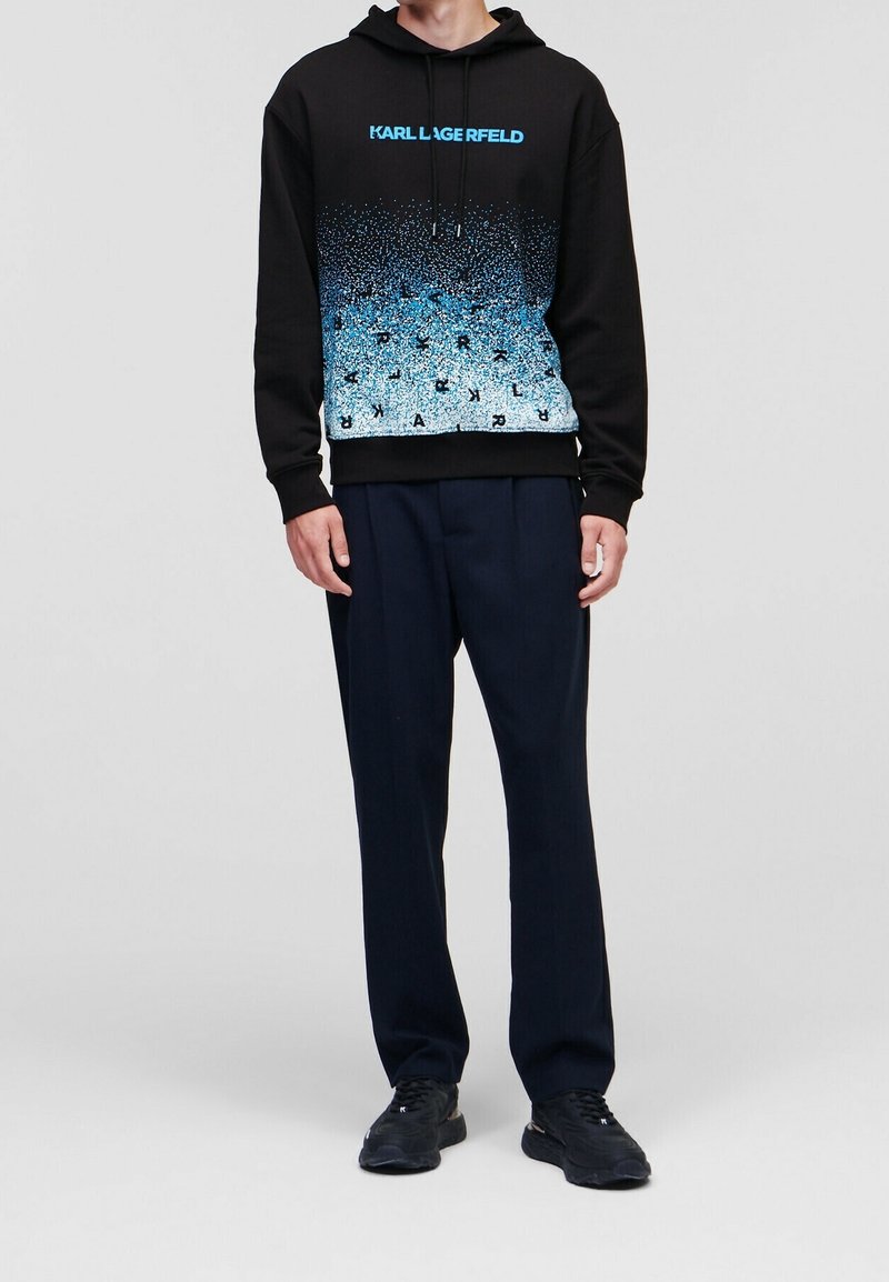 Homme portant un sweat à capuche noir avec un motif bleu moucheté et le texte « KARL LAGERFELD », associé à un pantalon bleu marine et des baskets noires, debout immobile.