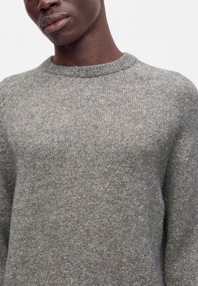 Gros plan d'une personne portant un pull tricoté gris doux avec un col rond sur fond blanc.