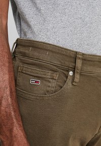 Pantalones vaqueros de mezclilla verde oliva con un bolsillo delantero que lleva un pequeño parche con logo. La tela tiene una textura suave con detalles de costuras visibles.