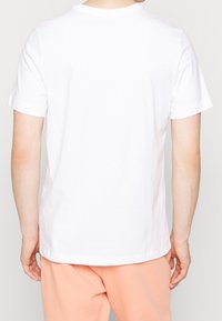 Henkilö, joka käyttää yksinkertaista valkoista lyhythihaista t-paitaa ja vaalean persikkaisia housuja, seisoo selkä kameraan päin.