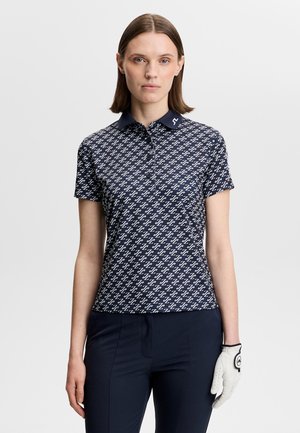 Donna che indossa una polo blu navy con motivo e pantaloni blu navy, con un guanto da golf bianco sulla mano sinistra, in piedi su uno sfondo chiaro e uniforme.