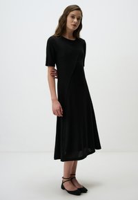 Robe noire mi-longue à manches courtes avec un motif texturé, présentant un design asymétrique. Ballerines noires avec des brides croisées.