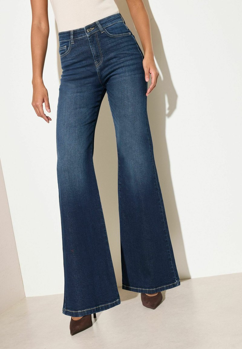 Žena má na sobě tmavě modré džíny s vysokým pasem a zvonovými nohavicemi, bílý nátělník bez rukávů a hnědé špičaté podpatky, stojí u světlé zdi.