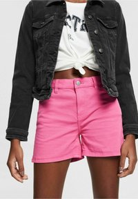 Musta farkkutakki etutaskuilla, yhdistettynä vaaleanpunaisiin puuvillahousuihin, joissa on nappikiinnitys ja suora leikkaus.