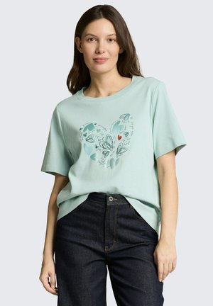 Vrouw draagt een lichtblauw t-shirt met een hartvormig ontwerp van blauwe en rode harten en zwarte krabbels, gecombineerd met een donkere spijkerbroek.