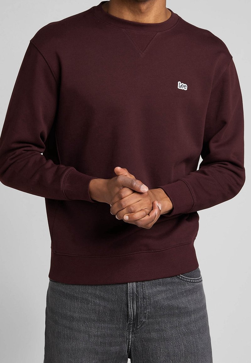 Personne portant un sweat-shirt Lee bordeaux foncé et un jean gris, mains jointes devant, vue des épaules aux hanches sur un fond neutre.