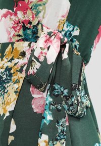 Vestido floral con una base verde, adornado con patrones en rosa, amarillo y azul verdoso. Presenta un detalle de cintura anudada con una textura suave.