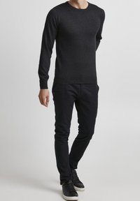 Dunkelgrauer Rundhals-Pullover aus strukturiertem Strickmaterial, kombiniert mit schmal geschnittenen schwarzen Hosen und schwarzen Sneakern, vor einem hellen Hintergrund stehend.