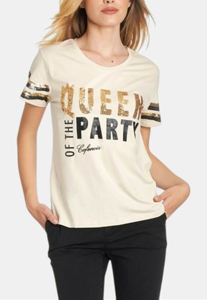 Donna che indossa una maglietta bianca con scritta dorata e nera "QUEEN OF THE PARTY" e pantaloni neri, mani nelle tasche.